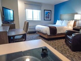 Hotel suite featuring a bedroom with a large bed and white bedding, a sitting area with a TV, and a view into the kitchenette.